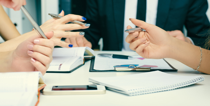 Group of 4 people at table with 3 women's hands holding pens above the table with notebooks, phone, and books on the table